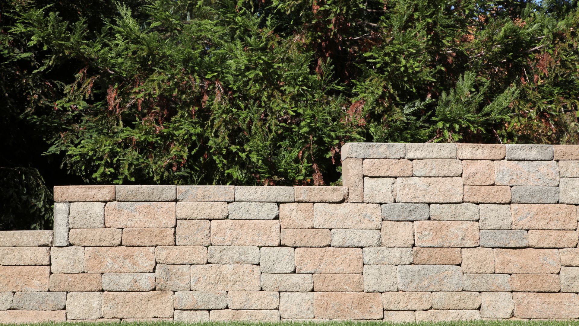 Stone retaining wall with textured blocks against lush green evergreen backdrop